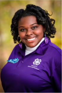 Portrait photo of a young woman dressed in a clean, purple polo featuring the 4-H logo. She has a bright smile and the photo is well lit with a simple and blurred photo. She has her hair well done with no hats or oversized jewelry. 
